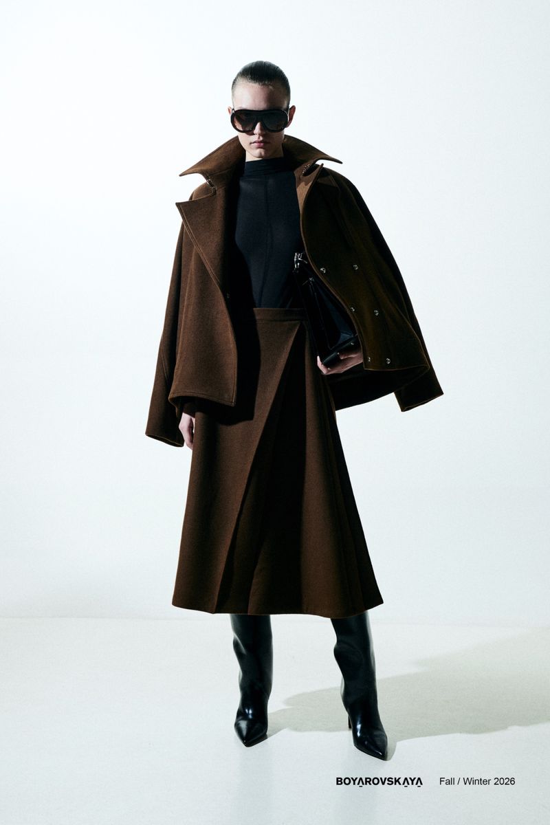 Fashion editorial shot of a female model in a brown coat, skirt, sunglasses, and black boots, posing in a bright photo studio.