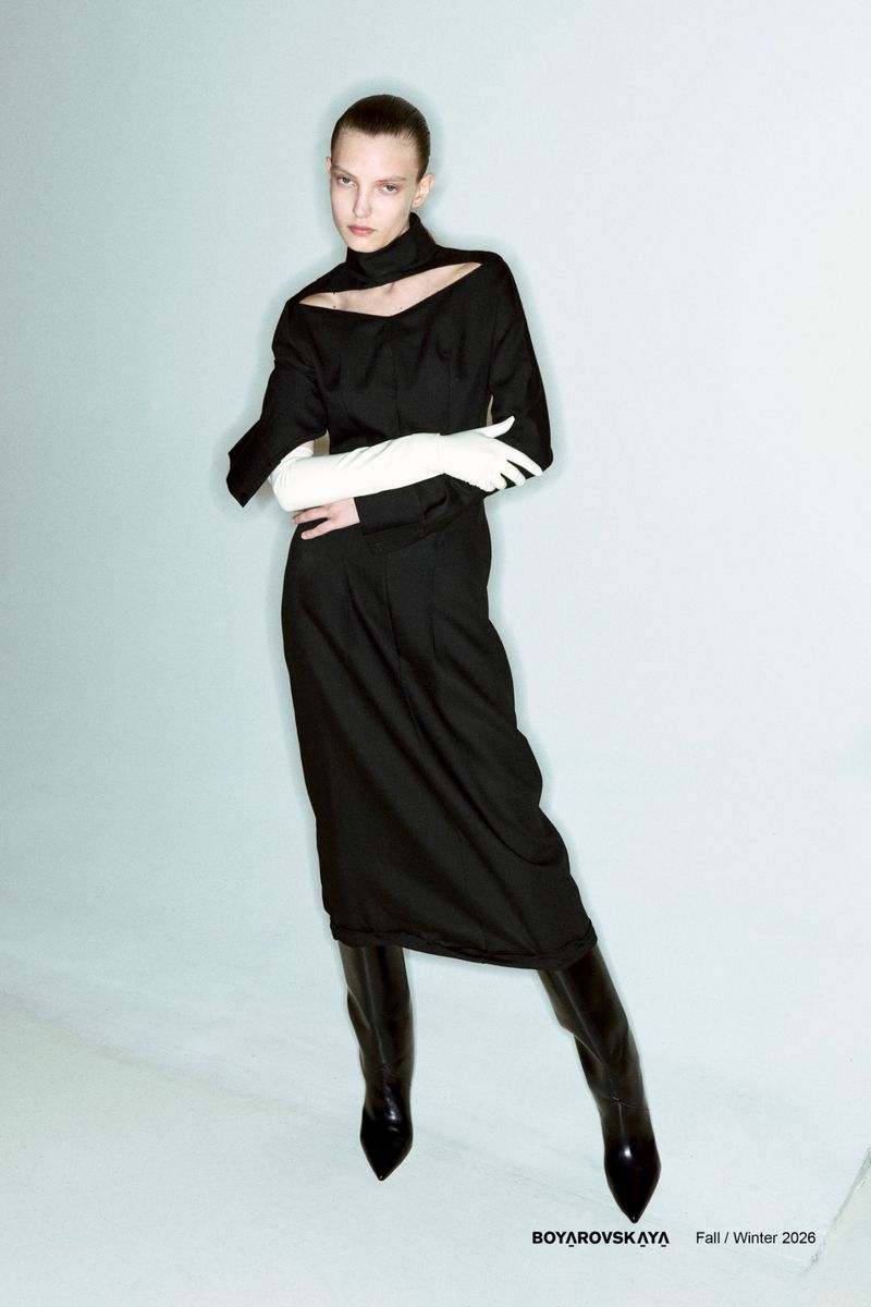 Fashion editorial shot of a young adult female model in a black dress and boots, wearing gloves, in a light blue studio setting.