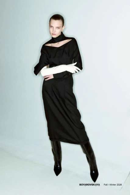 Fashion editorial shot of a young adult female model in a black dress and boots, wearing gloves, in a light blue studio setting.