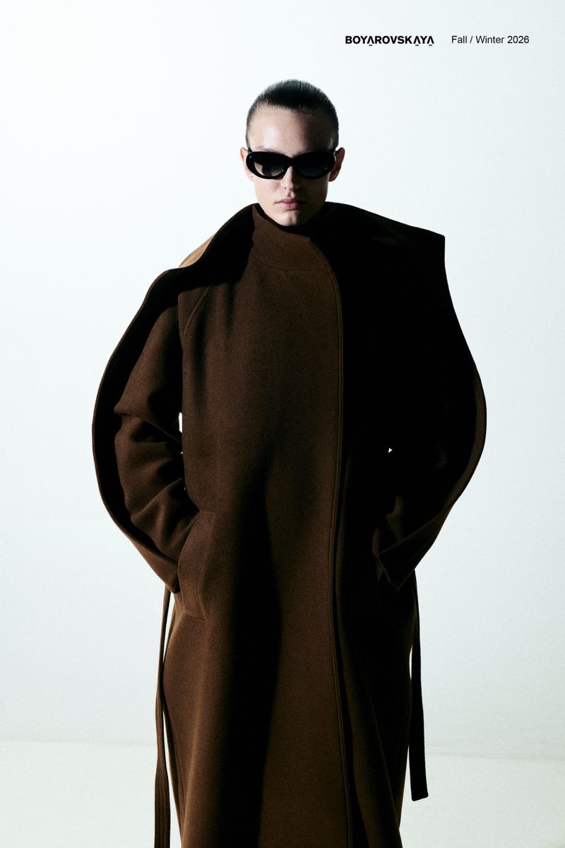 Fashion shot of adult female model with sunglasses wearing long, brown, belted coat against a clean, bright backdrop.