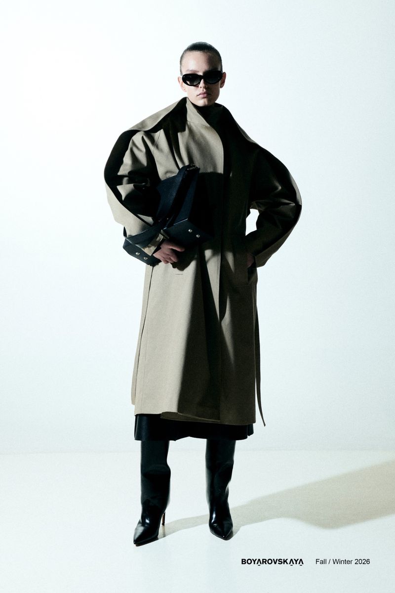 Model in dark sunglasses wearing an oversized beige trench coat and black boots holding a black handbag against a white background