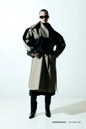 Model in dark sunglasses wearing an oversized beige trench coat and black boots holding a black handbag against a white background