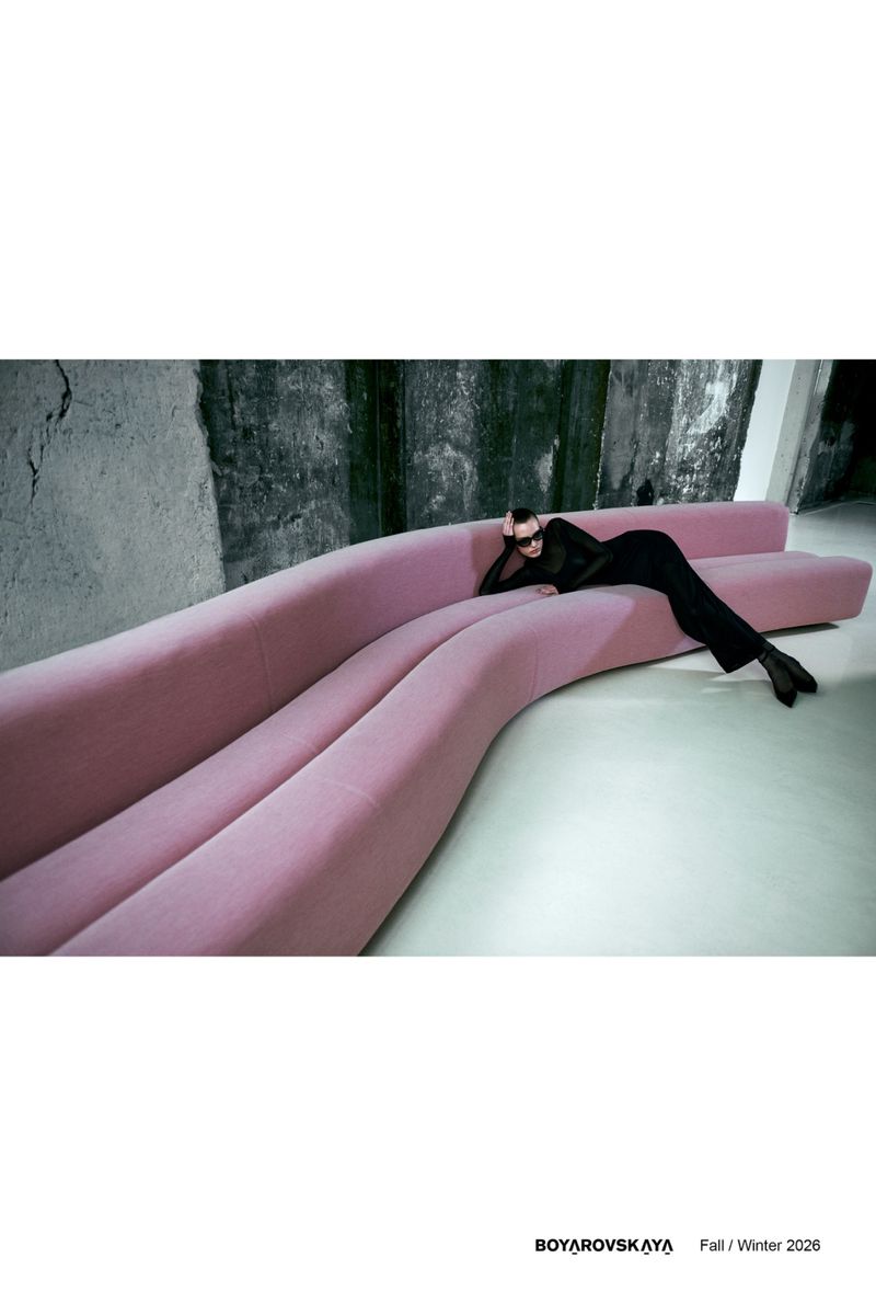 A woman in a black outfit lounges on a long, light pink couch against a gray concrete wall in an editorial fashion photography studio shot.
