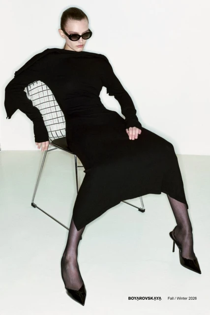 Elegant adult female fashion model in a long black dress, sheer hosiery, and heels sits on a metal chair against a white backdrop.