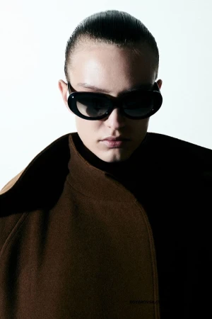 Model in dark sunglasses and brown collared coat against a plain white background