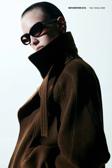 Fashion editorial featuring a woman in a brown coat and black sunglasses, posing indoors with a bright, neutral backdrop and diffused lighting.
