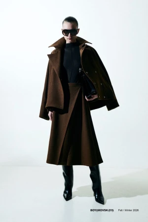 Model in dark sunglasses wearing a brown coat and matching skirt with black boots against a plain white background.