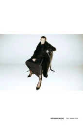 Model seated on a chair wearing a black coat and long skirt against a white studio background.