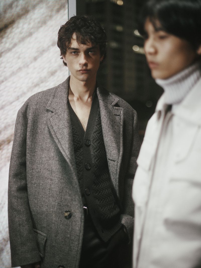 Two male models pose indoors wearing coordinated grey and black outfits, one in a coat and the other in a white jacket.