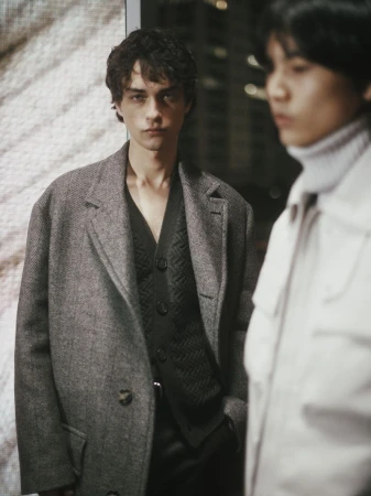 Two male models pose indoors wearing coordinated grey and black outfits, one in a coat and the other in a white jacket.