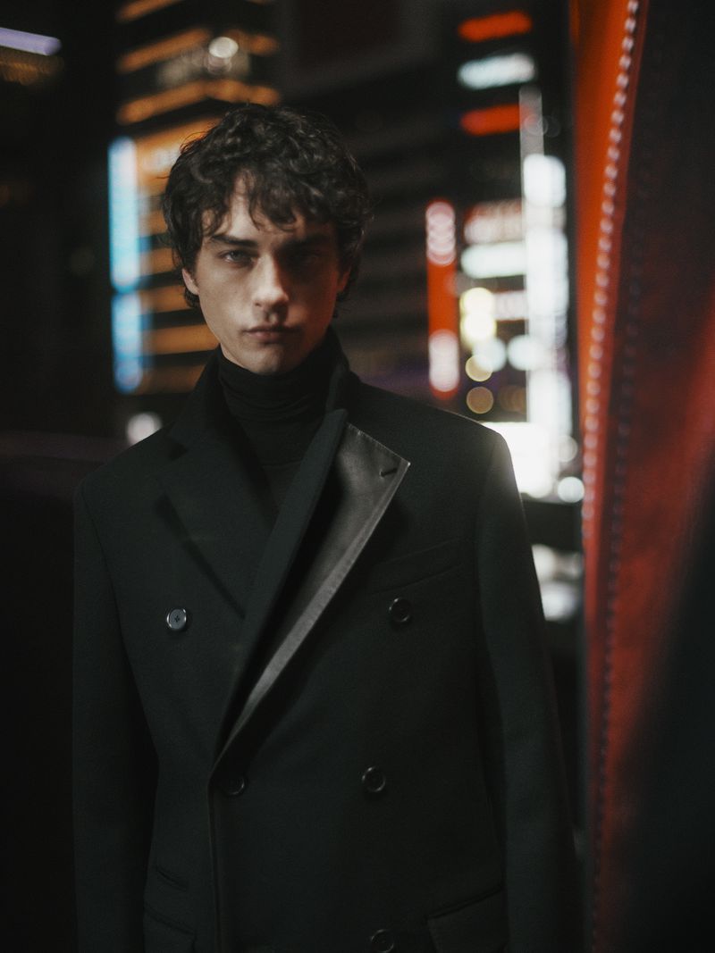 Portrait of a young adult man with curly hair wearing a dark overcoat and turtleneck against a blurred urban nightscape.