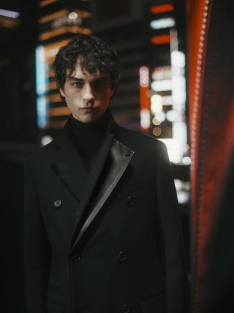 Portrait of a young adult man with curly hair wearing a dark overcoat and turtleneck against a blurred urban nightscape.