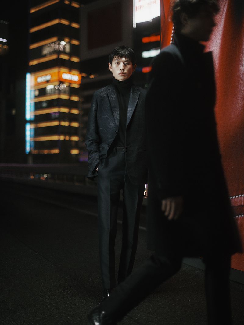Stylish man in a patterned suit poses on a city street at night with blurred background lights and a second man passing in front.