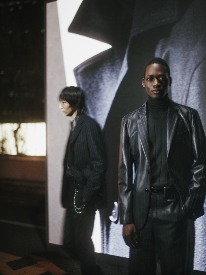 Two adult male models in pinstripe suits pose in front of a screen projection featuring a close-up of a gray wool coat.