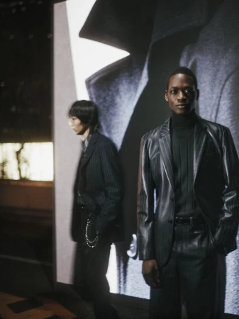 Two adult male models in pinstripe suits pose in front of a screen projection featuring a close-up of a gray wool coat.