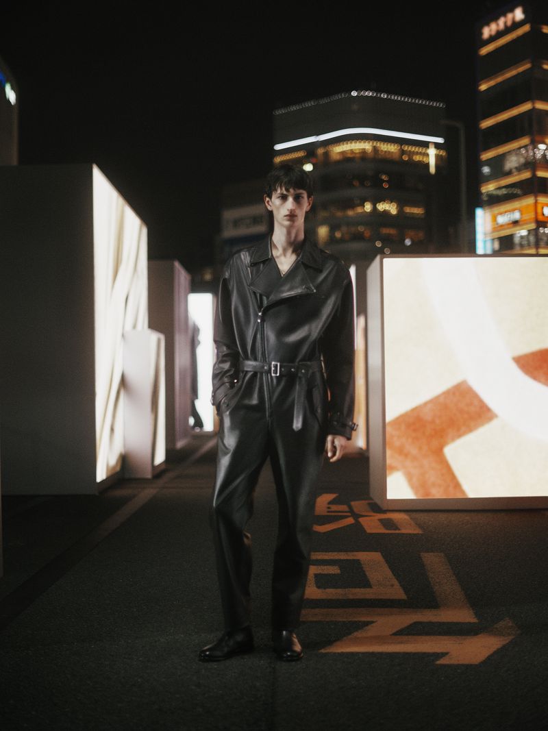 Male model in a black leather jumpsuit poses on a city street at night, surrounded by illuminated displays and city buildings in the background.
