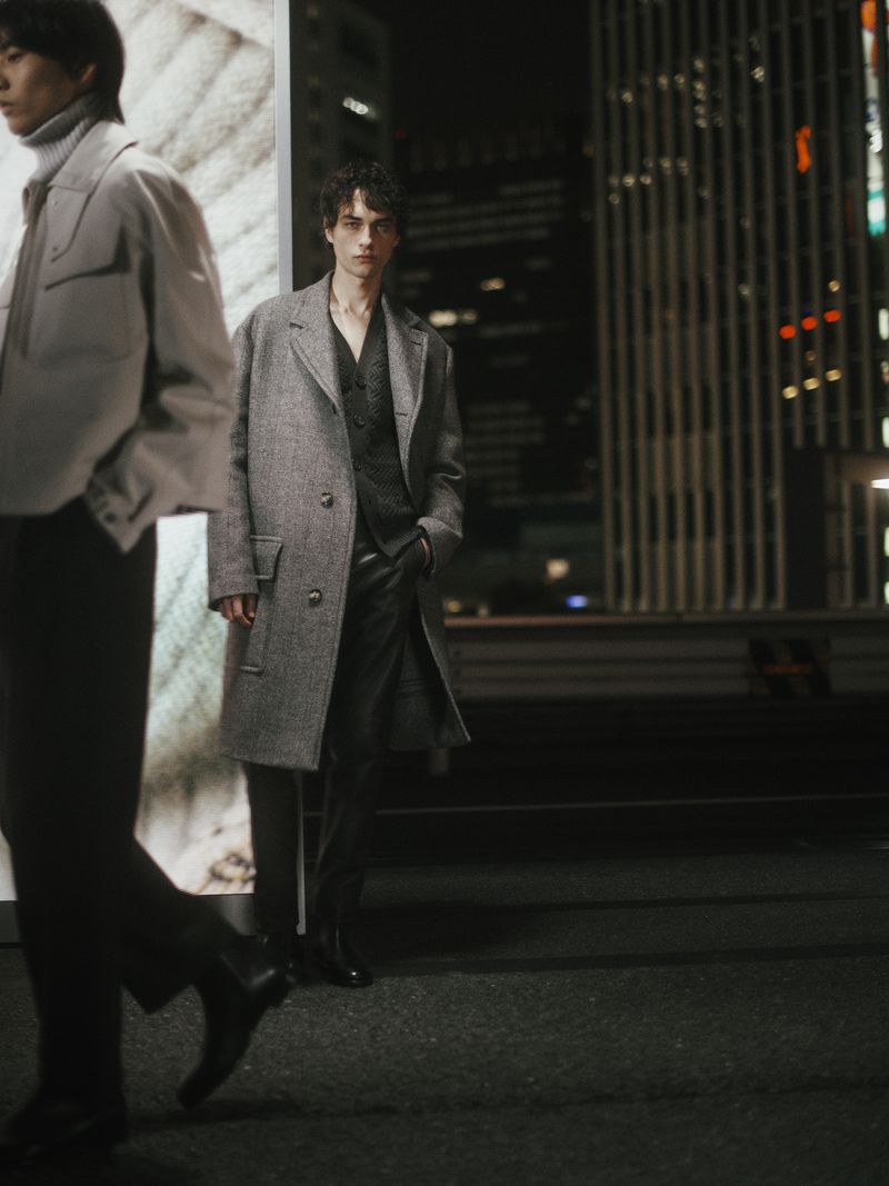 Two adult male fashion models in a city at night. One is walking and blurred, the other is posing in a wool coat.