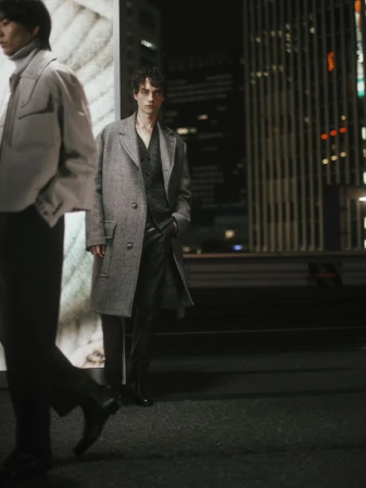 Two adult male fashion models in a city at night. One is walking and blurred, the other is posing in a wool coat.