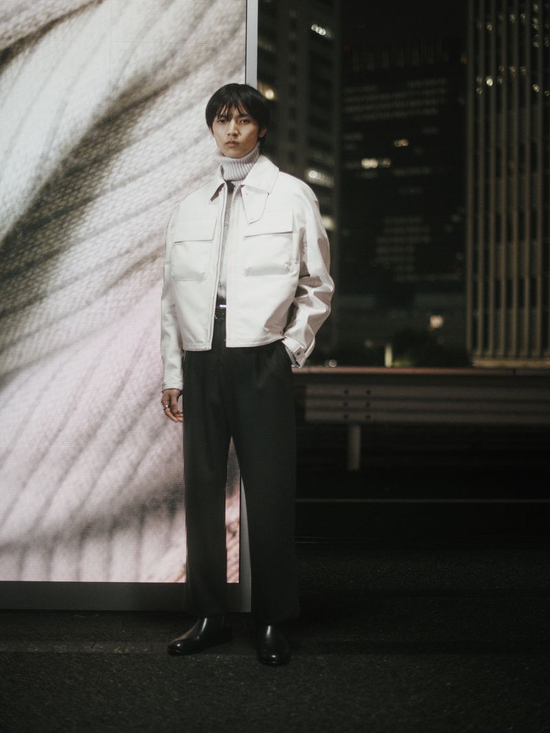 An Asian male model poses in a beige jacket, dark trousers, and black leather shoes against a backdrop of a cityscape at night.