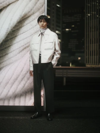 An Asian male model poses in a beige jacket, dark trousers, and black leather shoes against a backdrop of a cityscape at night.