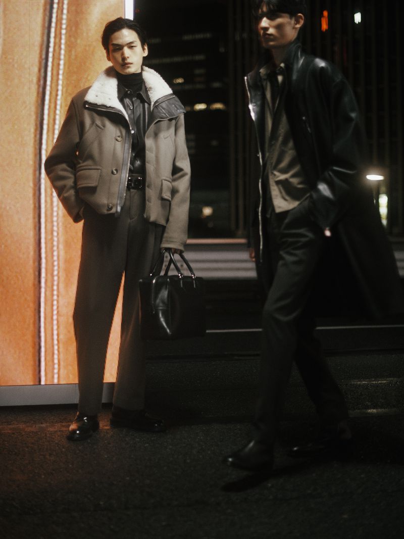 Two men pose on a city street at night, showcasing modern menswear. One wears a shearling jacket; the other, a long black coat.