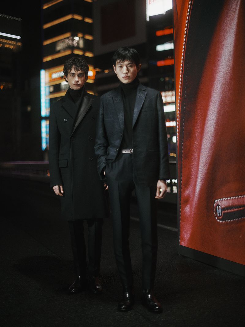 Two young men in dark suits pose in a city street with bright building lights and a red advertisement, fashionably styled in an urban setting.