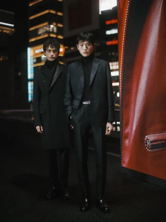Two young men in dark suits pose in a city street with bright building lights and a red advertisement, fashionably styled in an urban setting.