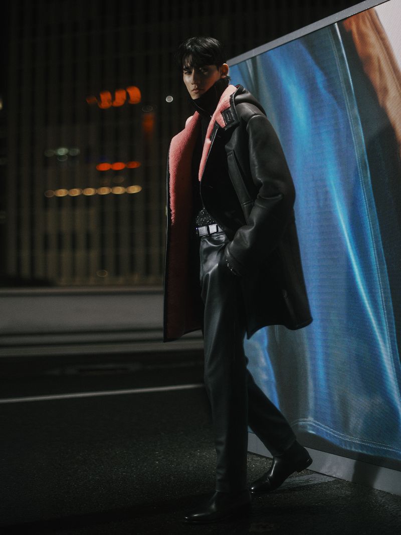 A stylish Asian male model poses in a black leather coat with pink lining and matching pants in an urban setting at night.