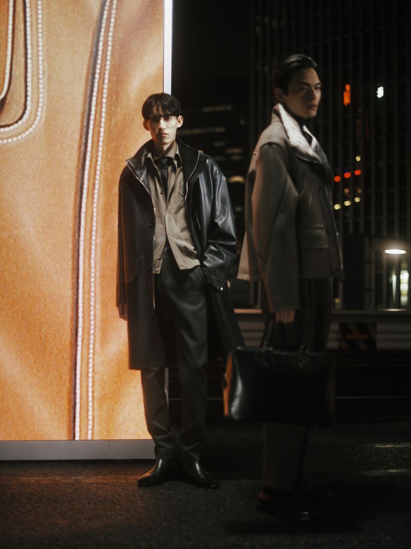 Two adult men pose in black coats in front of a large screen at night in an urban setting; one man holds a black briefcase.