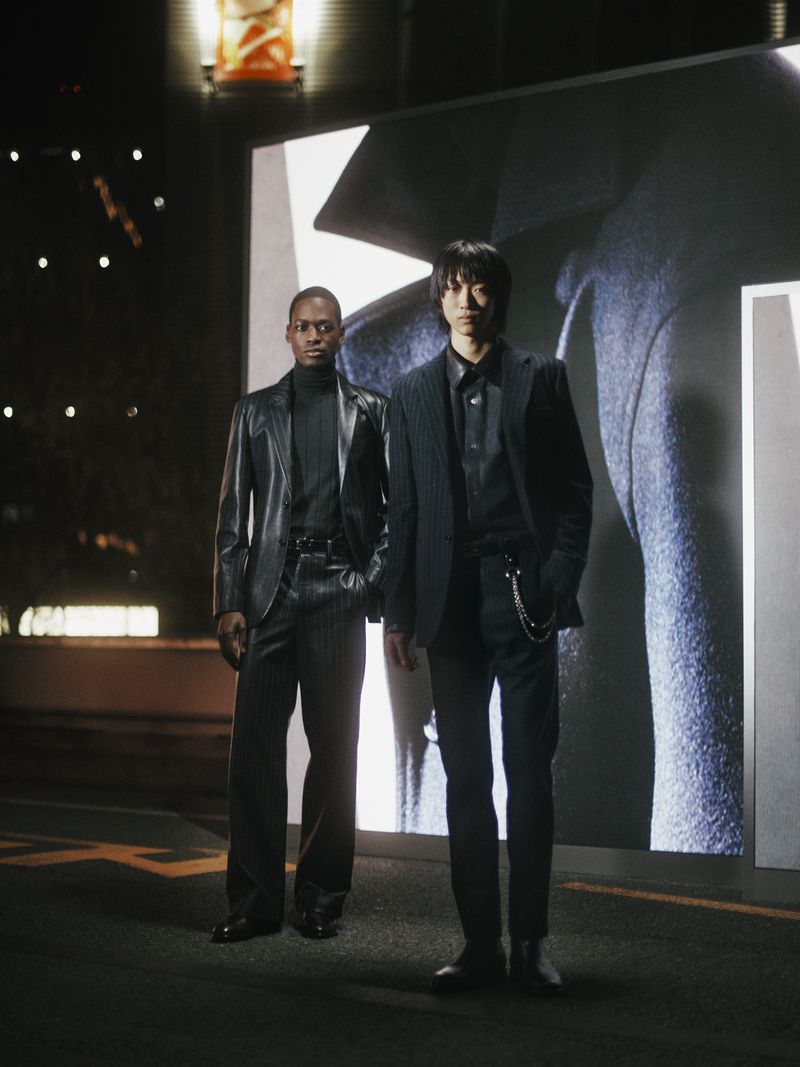 Two men in suits stand on a street at night in front of a digital billboard with an image of a coat, fashion photography.