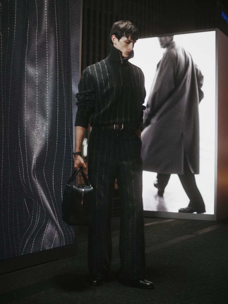 A full shot of a male fashion model showcasing a striped suit, standing in front of a light box with a similar image.