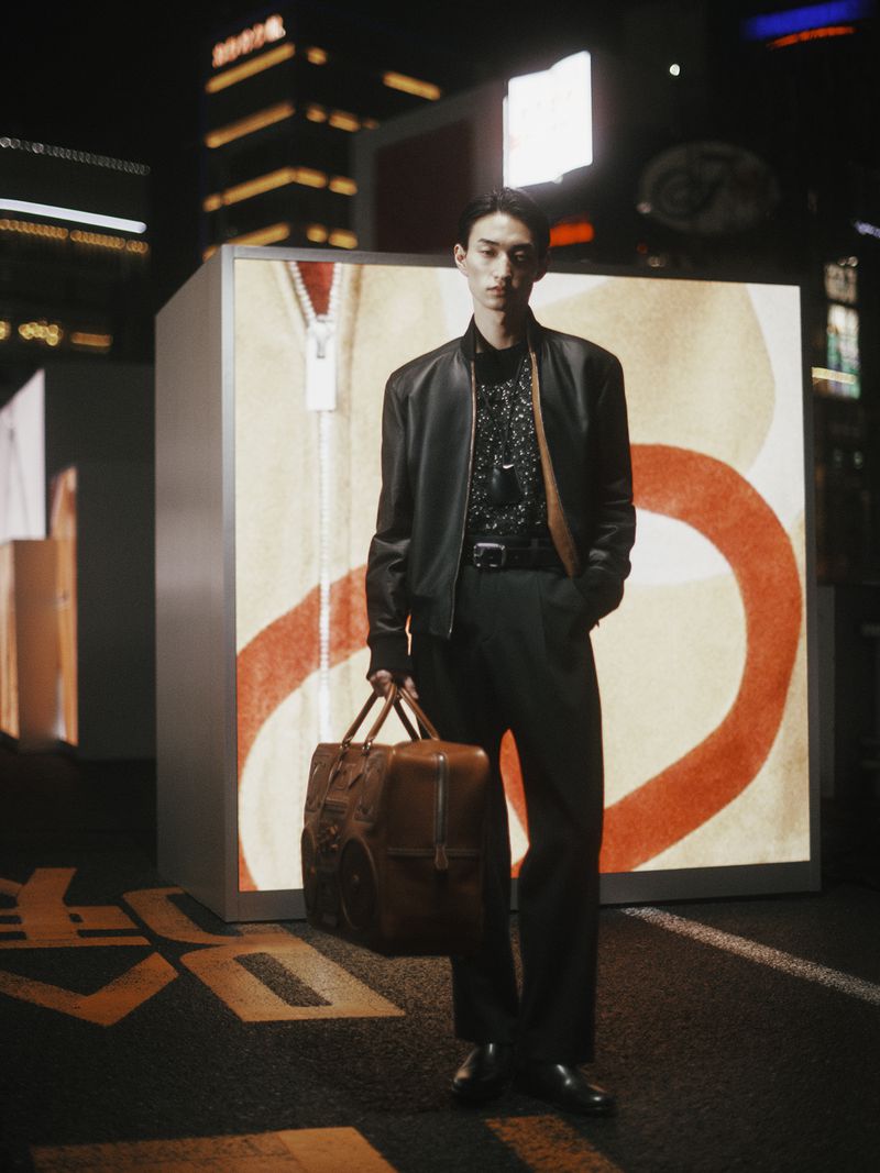 Asian man in a black jacket stands on a city street at night with a brown leather duffel bag, posing near illuminated artwork.