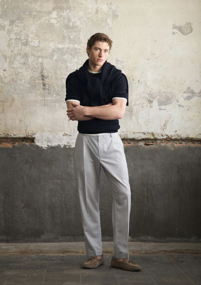 Full body studio shot of a fair-skinned, auburn-haired adult male wearing a dark blue short-sleeved sweater, gray trousers, and brown loafers.