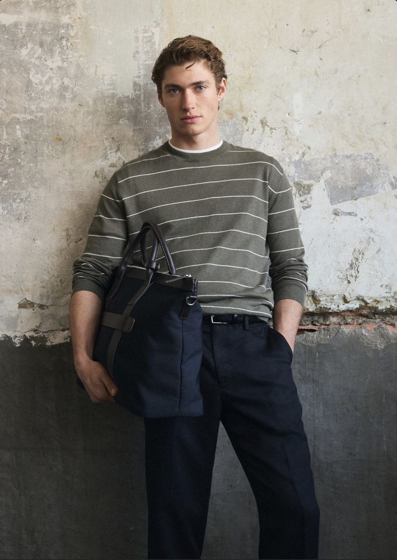 A stylish man wearing a striped sweater and dark pants holds a large bag against a textured wall, in a fashion portrait.