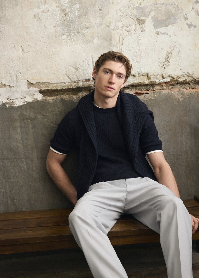 A fair-skinned adult male sits on a wooden bench in casual wear. He is dressed in a dark blue top with light grey pants.
