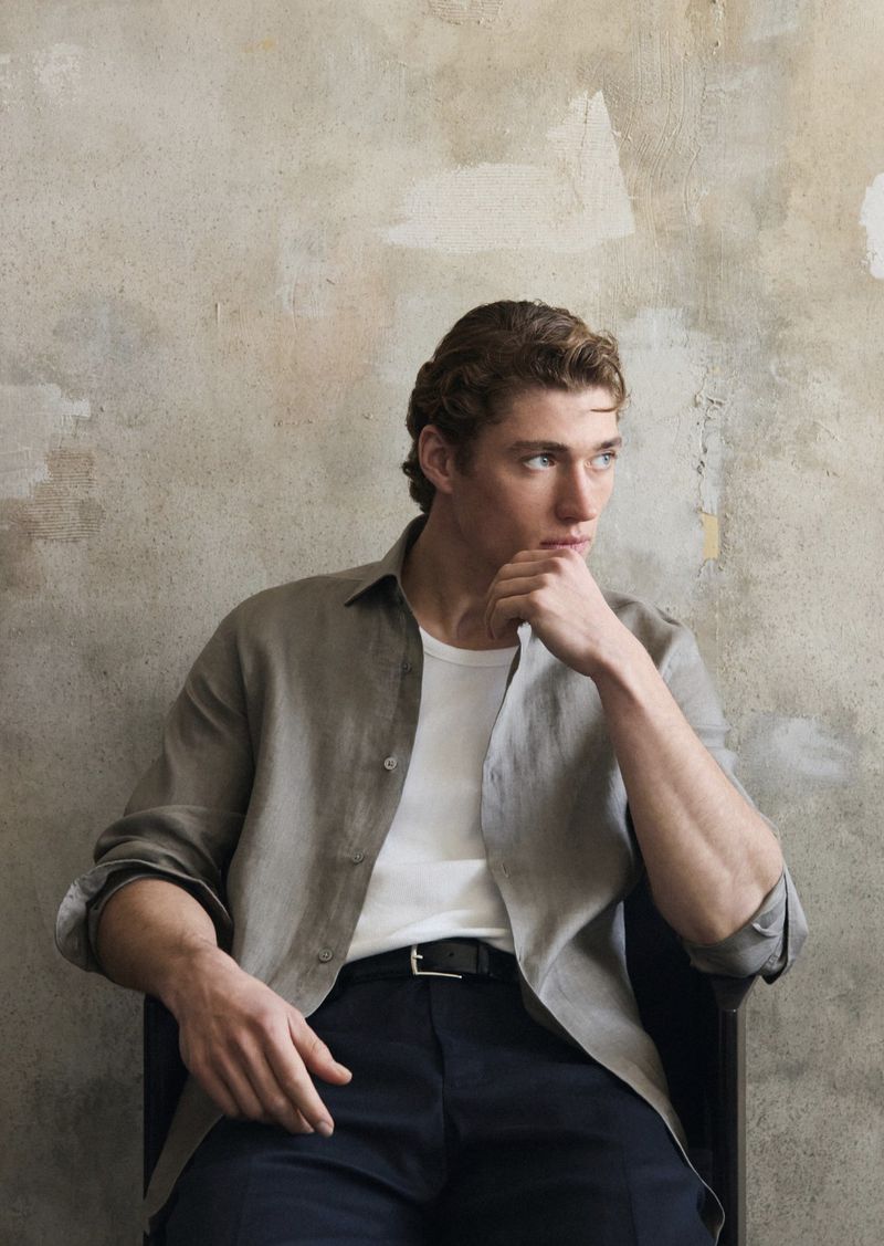 Adult male in a casual light brown button-down shirt and dark pants, sitting in a chair against a textured grey studio backdrop.