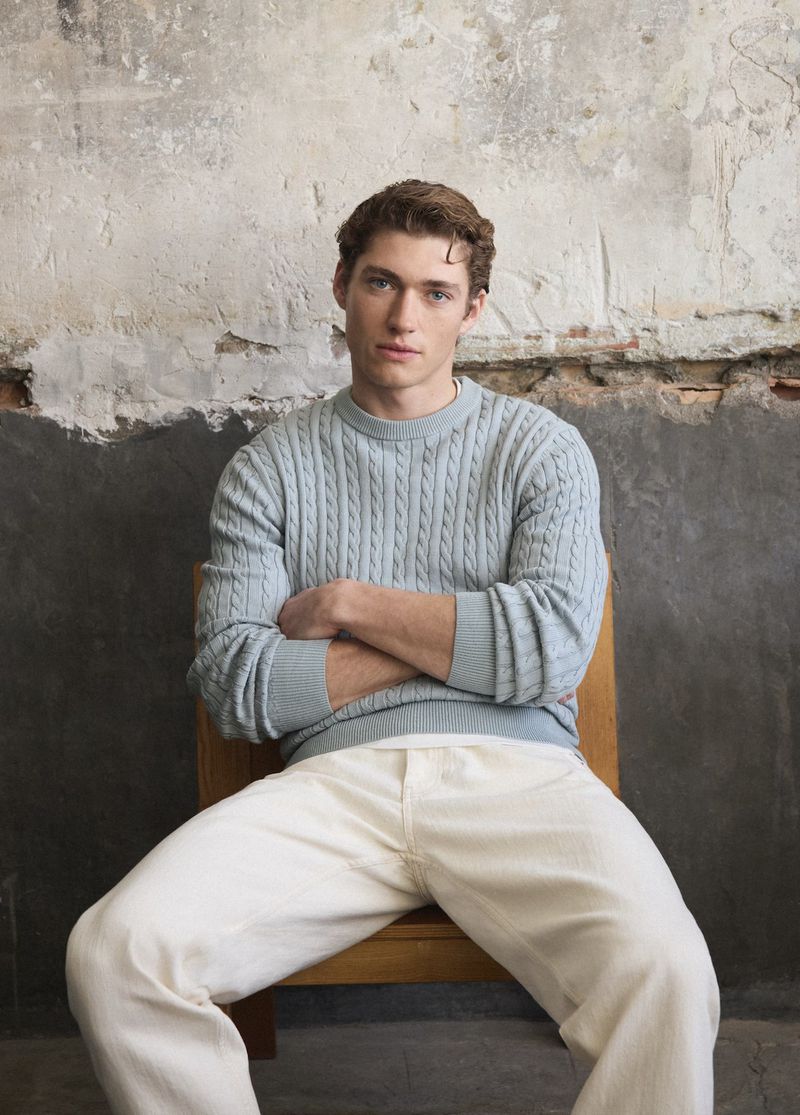 Indoor portrait of a young adult male in a light blue sweater and white pants sitting on a wooden chair in front of a textured wall.