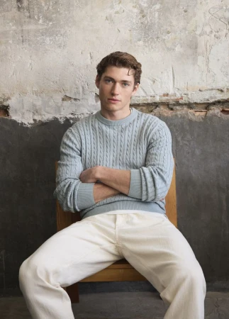 Indoor portrait of a young adult male in a light blue sweater and white pants sitting on a wooden chair in front of a textured wall.