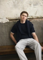 Young male model seated on a wooden bench wearing a dark knit sweater and light grey trousers against a textured wall.