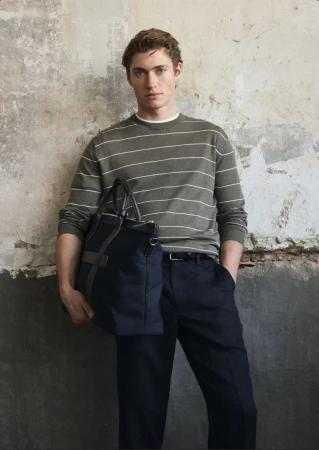 Man wearing a striped green sweater and dark trousers holding a dark blue tote bag against a weathered textured wall