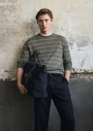 Man wearing a striped green sweater and dark trousers holding a dark blue tote bag against a weathered textured wall