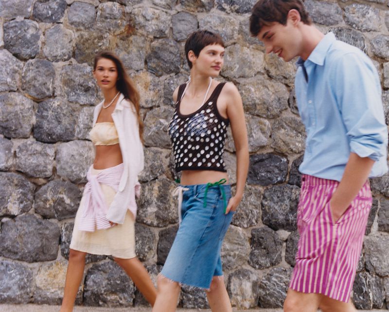 Three fashion models walk in front of a stone wall, showcasing summer attire: a tube top, tank top, shorts, and button-down shirt.