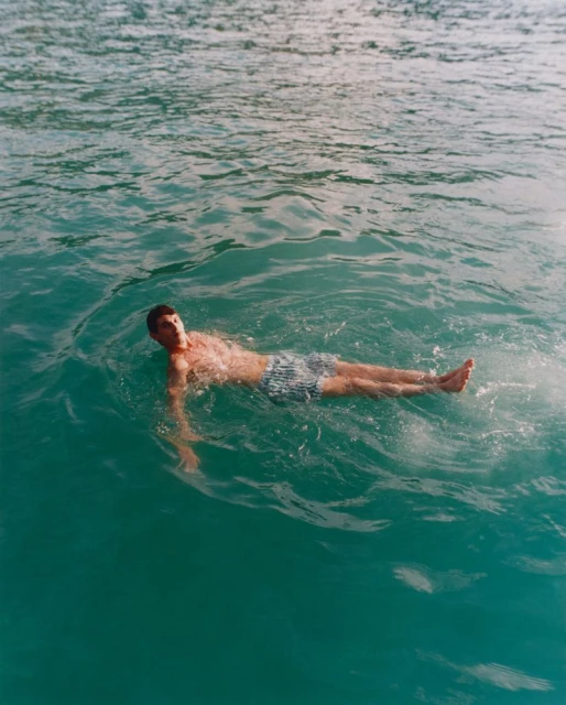 Man in blue and white striped swimming trunks floating on his back in teal-colored water on a bright, sunny day in an outdoor setting.