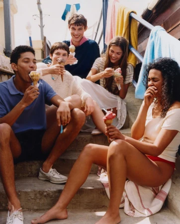 Five young adults sit on steps outdoors eating ice cream cones, with towels hanging in the background, creating a casual, summery scene.