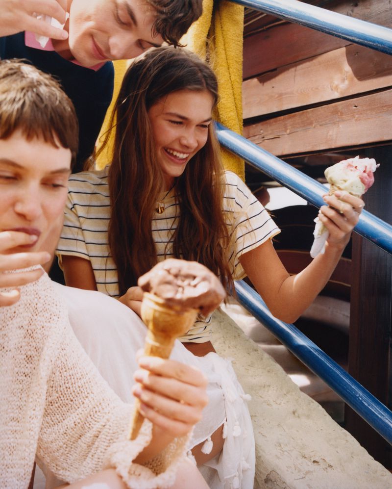 Three young adult friends smiling and holding ice cream cones on stairs; one female in striped top, two males in sweaters, outdoor setting.