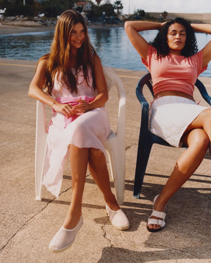 Two women in summer dresses are sitting in plastic chairs near the ocean, enjoying the sunlight and relaxing with a serene coastal view.