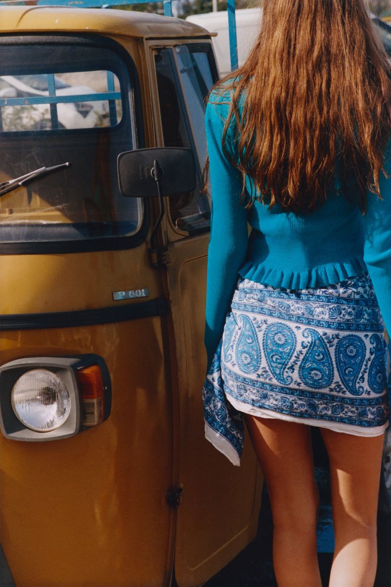 Young woman with long brown hair wearing a blue top and patterned sarong stands near a yellow three-wheeled vehicle outdoors.