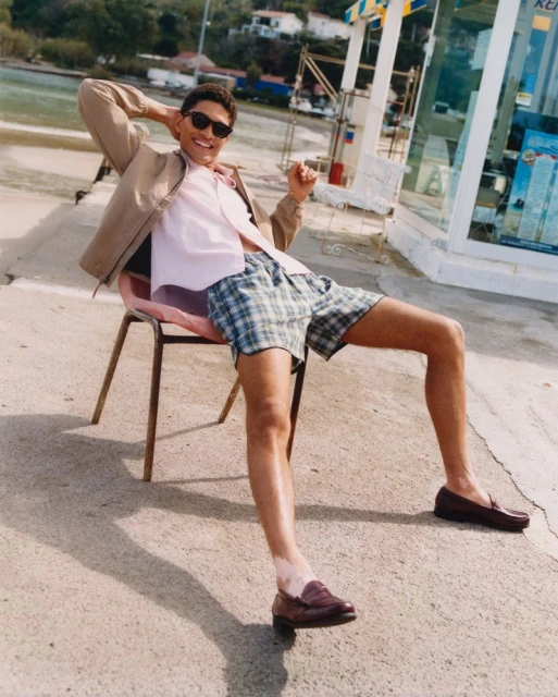 A man in a pink shirt and plaid shorts sits on a chair outside near a roadside kiosk on a sunny day with ocean in the background.
