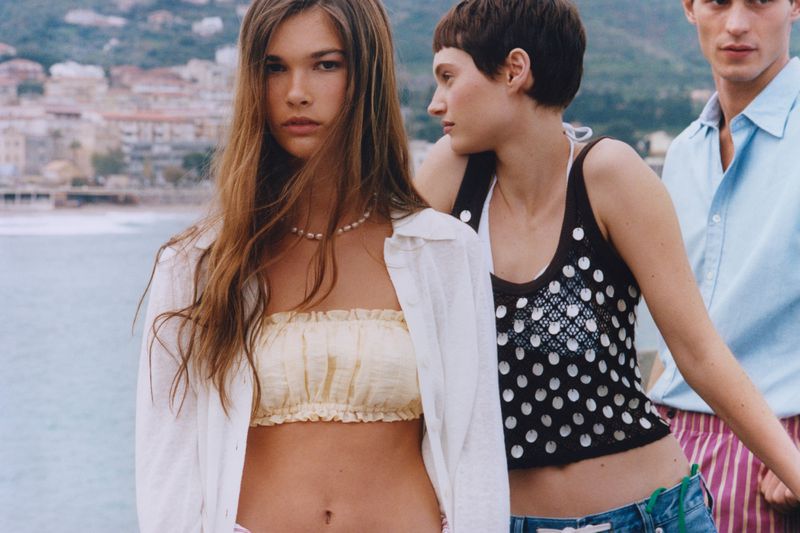 Three models pose outdoors with the sea and buildings blurred in the background, showcasing fashion and summer vibes.