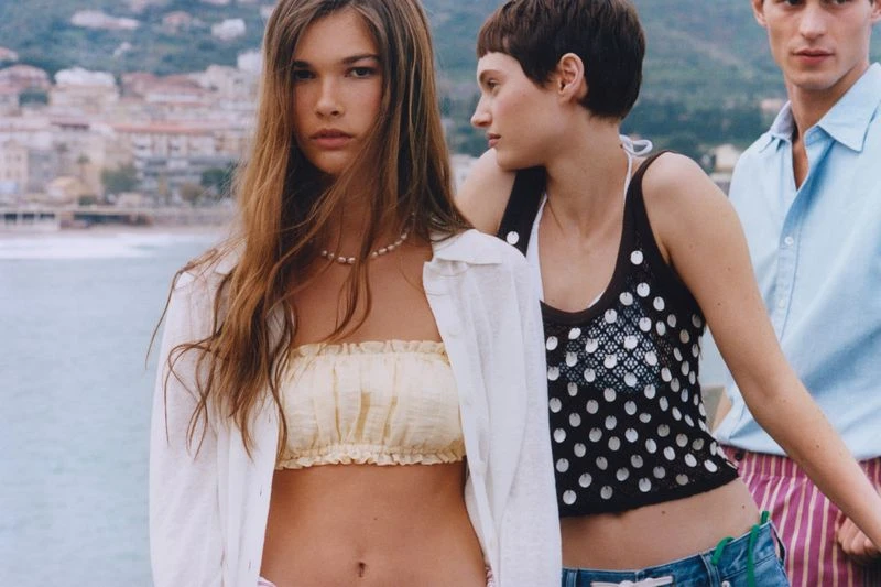 Three models pose outdoors with the sea and buildings blurred in the background, showcasing fashion and summer vibes.